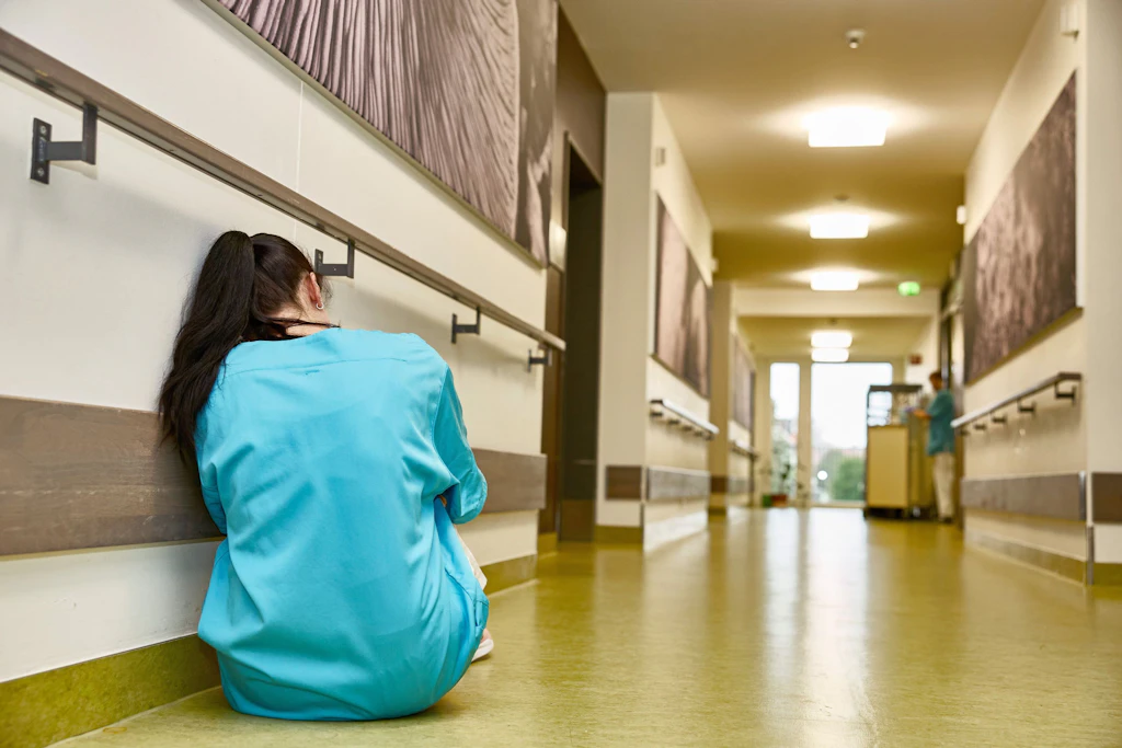 System am Limit: Eine erschöpfte Pflegekraft kauert auf einem Klinikflur.