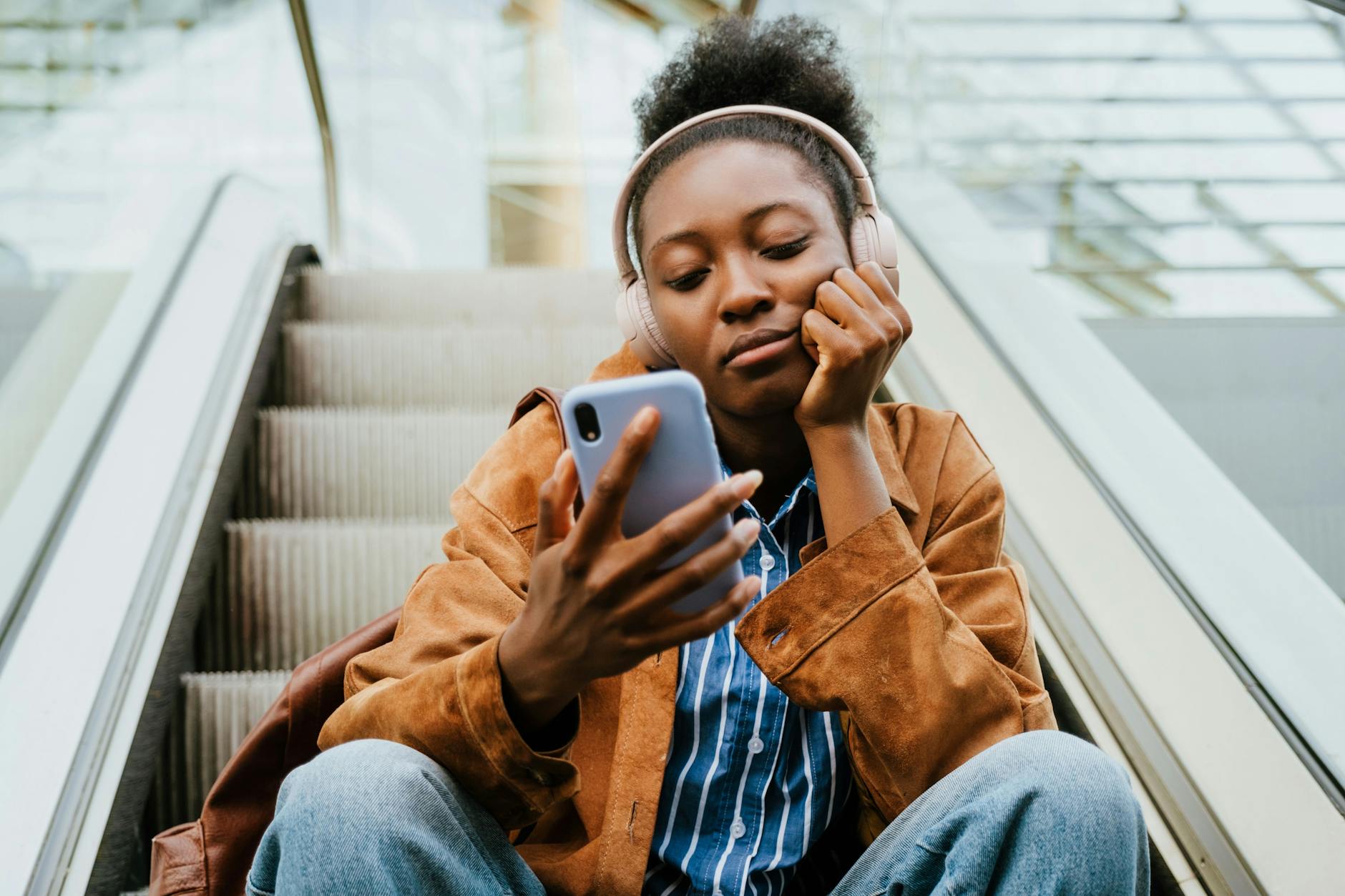 Young african american woman listening to music on headphones and using cellphone on escalator stairs