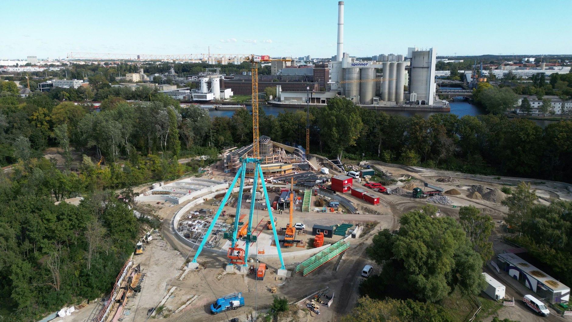 Blick aus der Drohnenperspektive auf die Baustelle, die bereits aufgestellten blauen Stützen des neuen Riesenrades und die Umrandung des Wasserbassins. Im Hintergrund, kurz vor der Spree und dem schmalen Waldstreifen, sieht man das neue Englische Dorf, an dem gerade auch gebaut wird.
