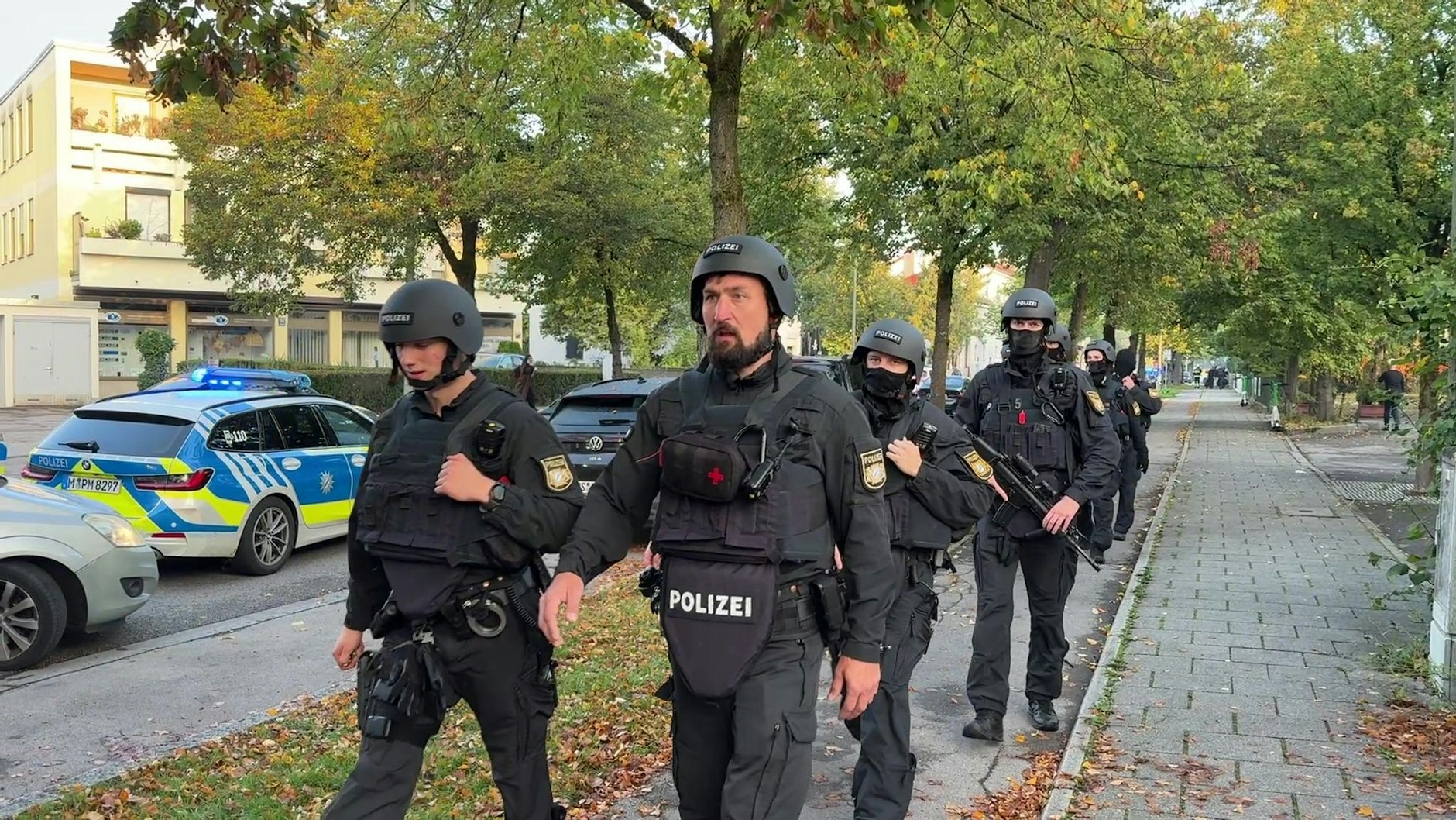 Polizisten mit Schutzwesten gehen über einen Fußweg (Screenshot aus einem Video). Ein Brand in einem Münchner Einfamilienhaus hat einen Großeinsatz von Polizei und Feuerwehr ausgelöst.