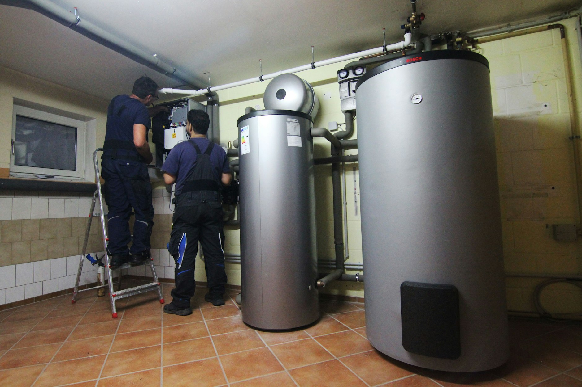 Zwei Handwerker der Firma Jänichen Versorgungstechnik stellen eine gerade installierte Wärmepumpe im Keller einer Doppelhaushälfte ein. (Symbolbild)