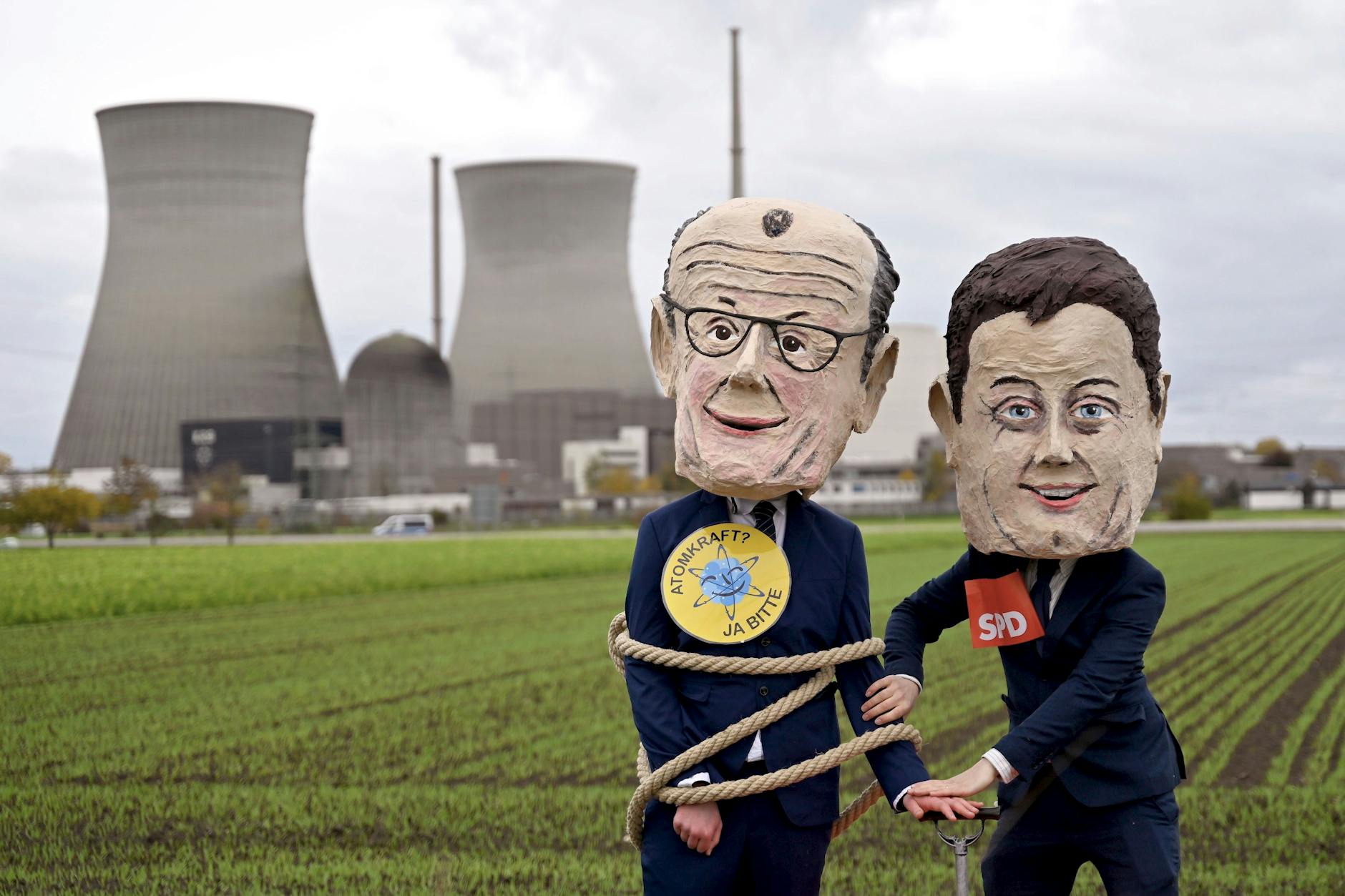 Das Atomkraftwerk im bayerischen Gundremmingen vor seiner Sprengung Ende Oktober: Demonstranten posieren mit Masken von Bundeskanzler Friedrich Merz und Vizekanzler Lars Klingbeil.