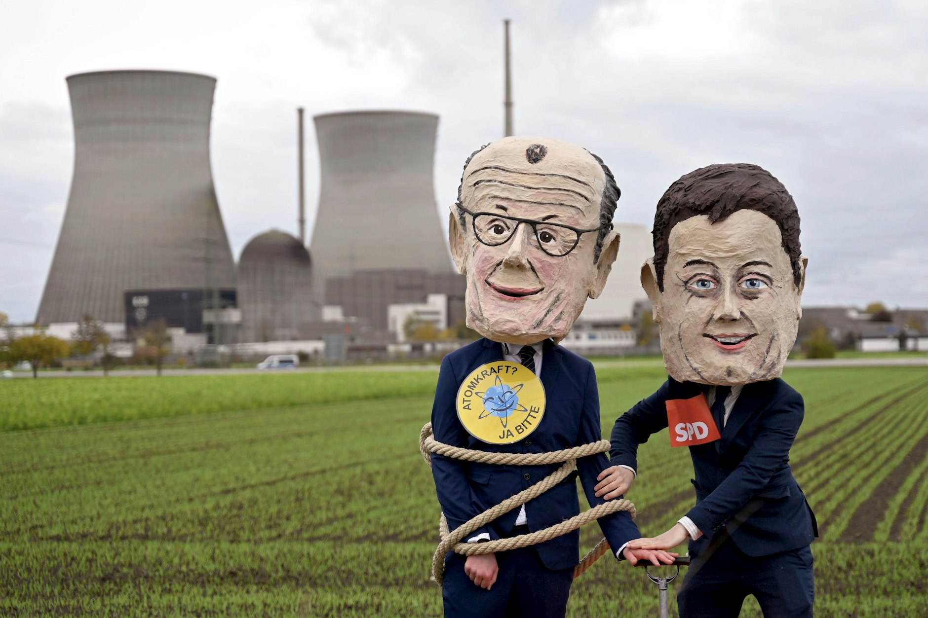 Das Atomkraftwerk im bayerischen Gundremmingen vor seiner Sprengung Ende Oktober: Demonstranten posieren mit Masken von Bundeskanzler Friedrich Merz und Vizekanzler Lars Klingbeil.