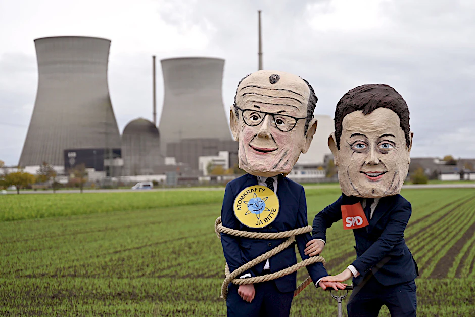 Das Atomkraftwerk im bayerischen Gundremmingen vor seiner Sprengung Ende Oktober: Demonstranten posieren mit Masken von Bundeskanzler Friedrich Merz und Vizekanzler Lars Klingbeil.