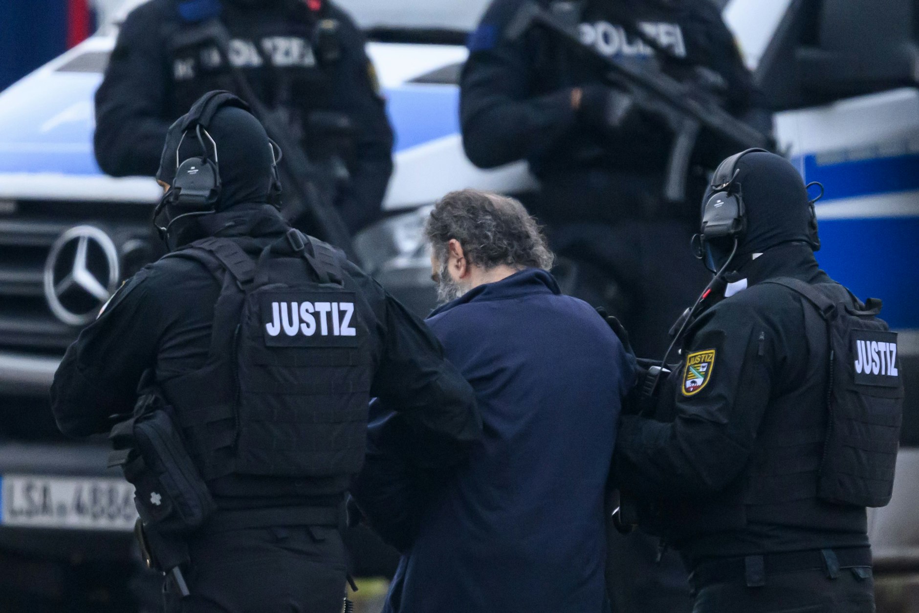 Der Angeklagte Taleb A. wird von maskierten Justizbeamten in den Gerichtssaal gebracht. Er ist an Händen und Füßen gefesselt.