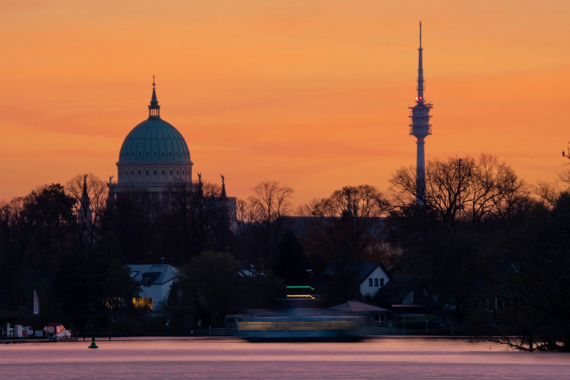 Hinter der St. Nikolaikirche in Potsdam und dem Fernmeldeturm Berlin-Schäferberg geht die Sonne auf, während eine Fähre die Havel überquert.