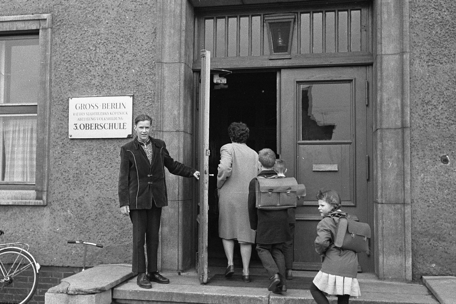 Schüler mit Tornister gehen mit einer Lehrerin in eine Schule.