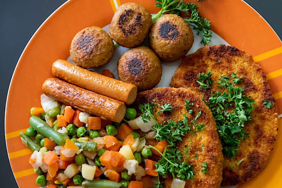 Vegane Falafel, vegane Schnitzel, vegane Würstchen und Gemüse sind auf einem Teller in einer Küche zu sehen.