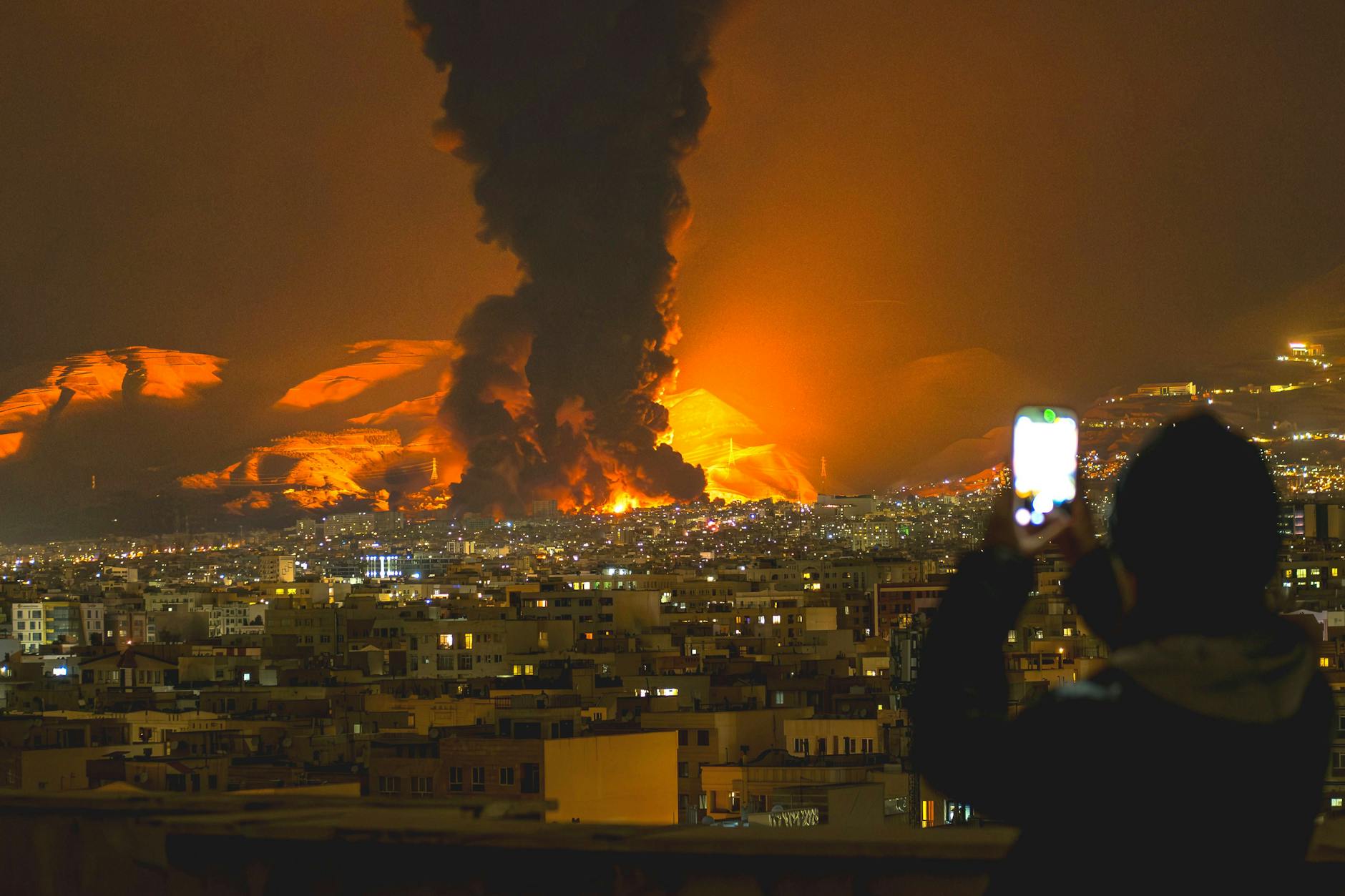 Eine Frau fotografiert mit ihrem Handy, während Rauch nach Luftangriffen auf ein Öldepot in Teheran aufsteigt.