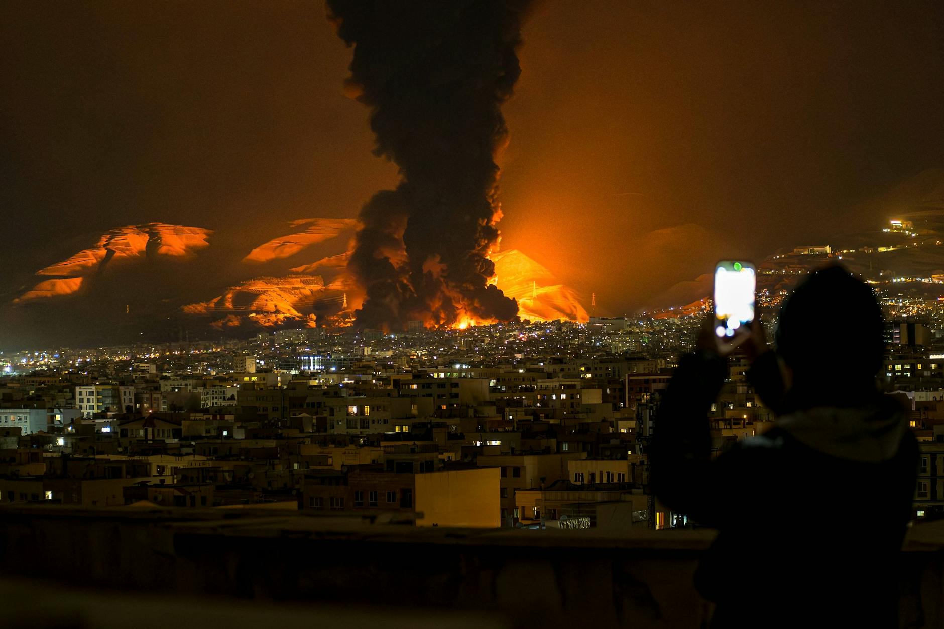 Eine Frau filmt mit ihrem Handy, wie am 7. März 2026 nach Luftangriffen auf ein Öldepot in Teheran Rauch und Flammen aufsteigen.