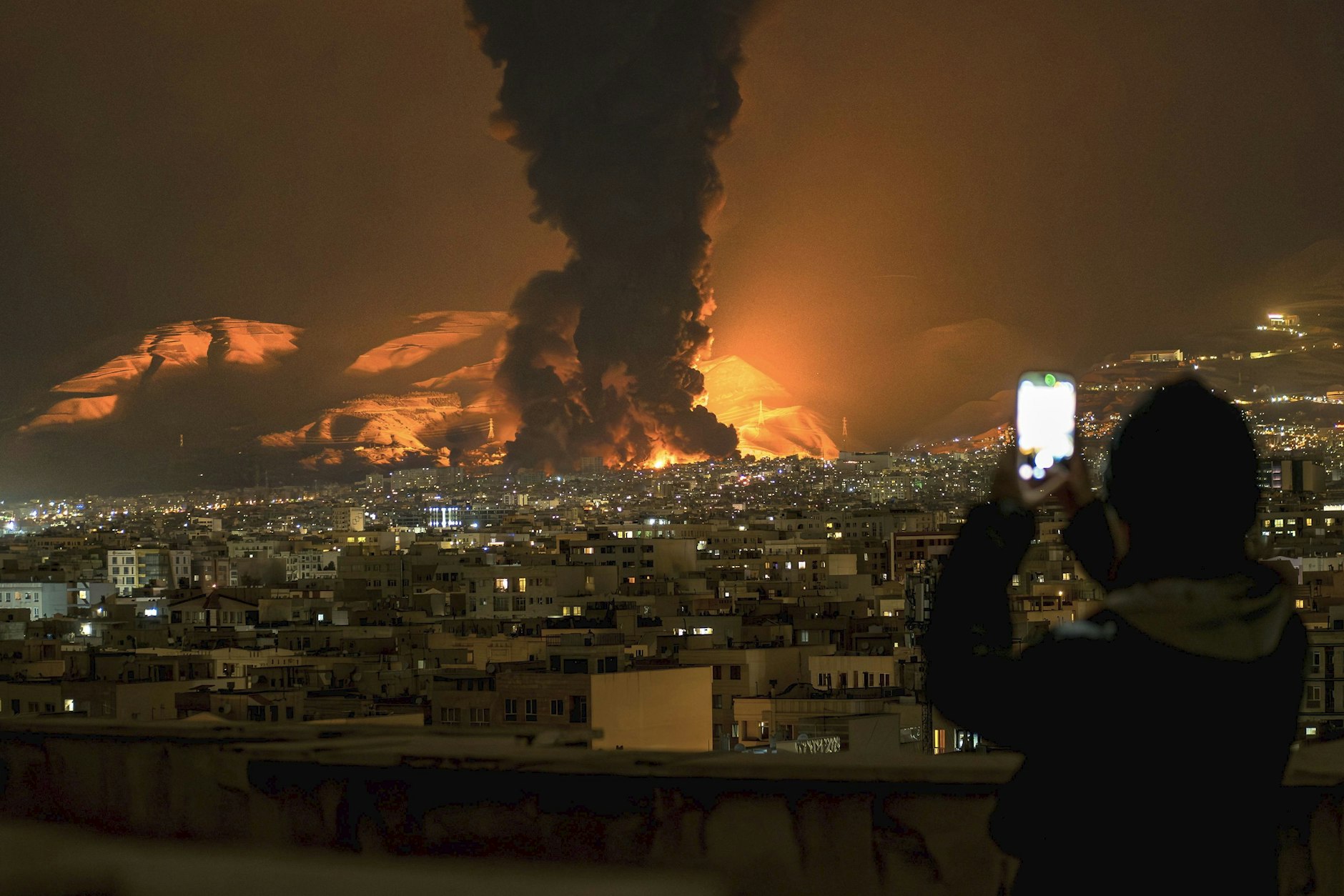 Eine Frau filmt mit ihrem Handy, wie am 7. März 2026 nach Luftangriffen auf ein Öldepot in Teheran Rauch und Flammen aufsteigen.