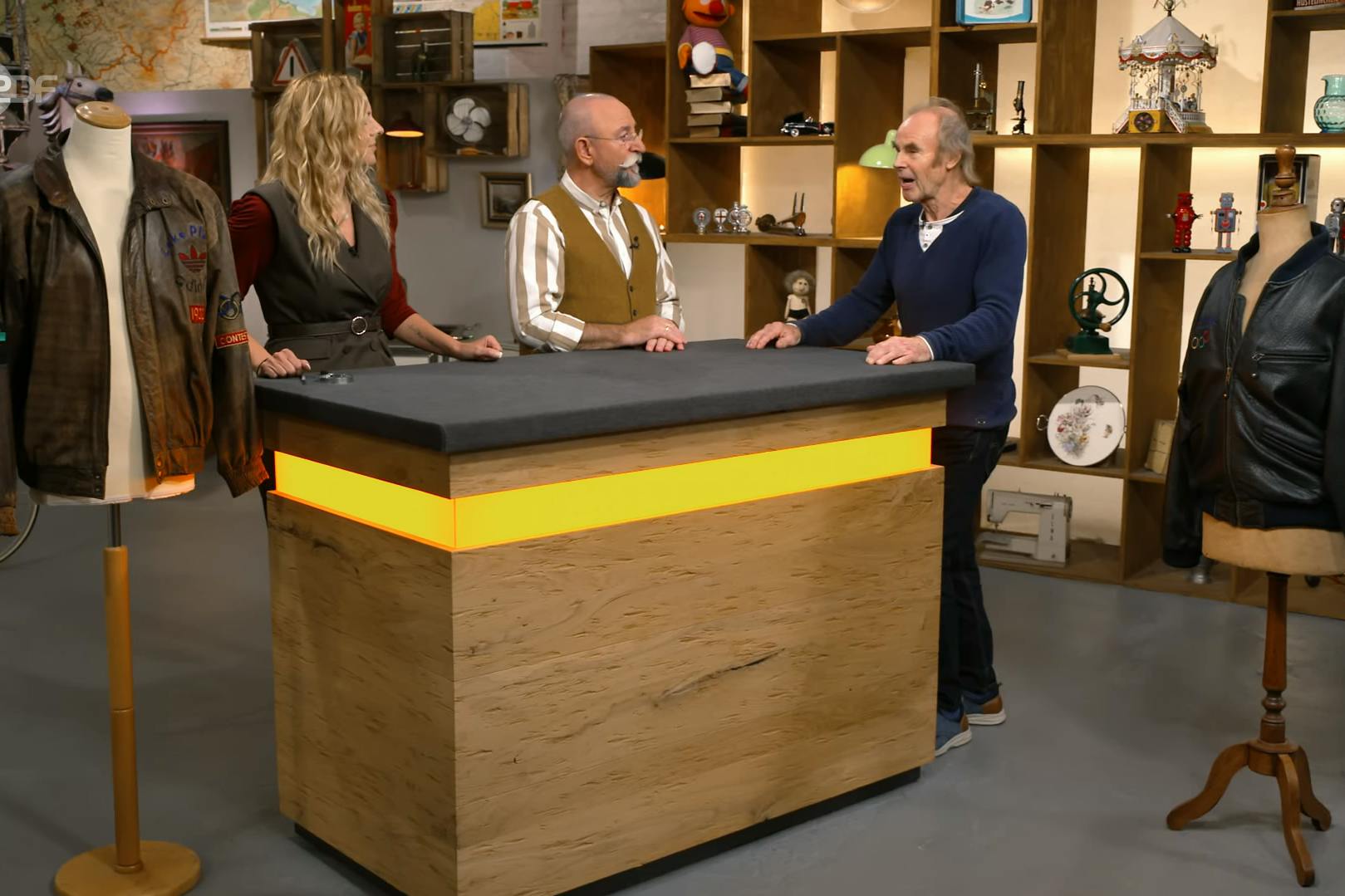 Mode-Expertin Annika Raßbach, Moderator Horst Lichter und Kandidat Anton Strauss (r.) schauen sich bei der Expertise bei „Bares für Rares“ die Lederjacken ganz genau an.