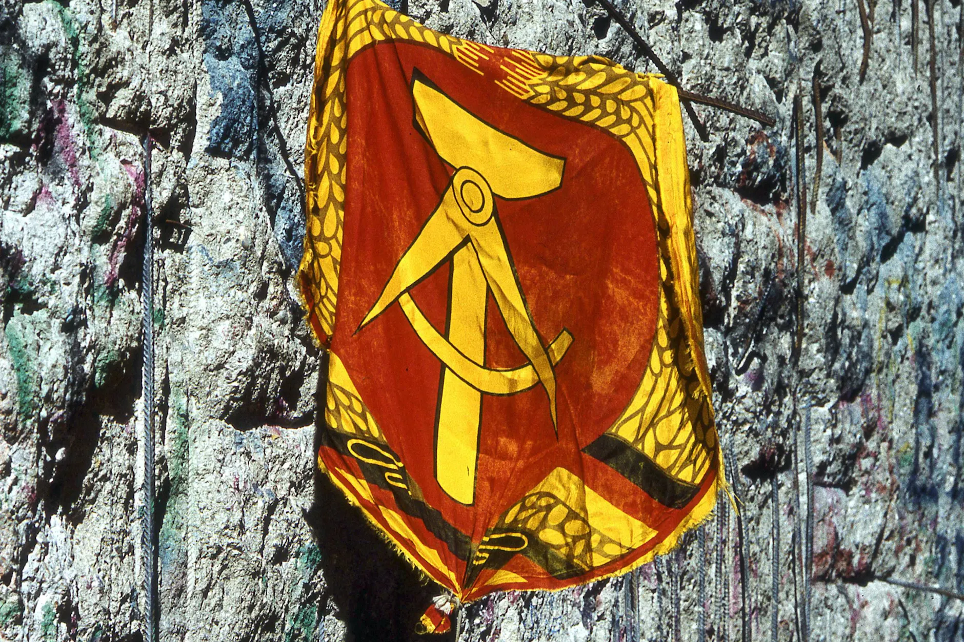 Vergangen, aber nicht vergessen: Das Wappen der DDR zeigt Hammer, Zirkel, Ährenkranz. Friedfertige Symbole. Im Hintergrund ein Stück der von Mauerspechten zerpickten Mauer