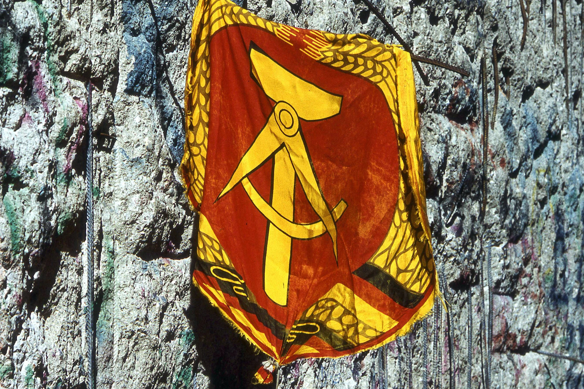 Vergangen, aber nicht vergessen: Das Wappen der DDR zeigt Hammer, Zirkel, Ährenkranz. Friedfertige Symbole. Im Hintergrund ein Stück der von Mauerspechten zerpickten Mauer