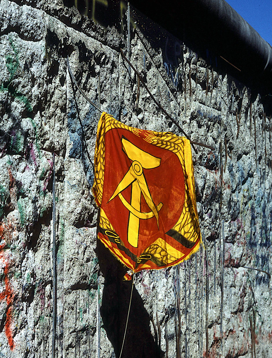 Vergangen, aber nicht vergessen: Das Wappen der DDR zeigt Hammer, Zirkel, Ährenkranz. Friedfertige Symbole. Im Hintergrund ein Stück der von Mauerspechten zerpickten Mauer