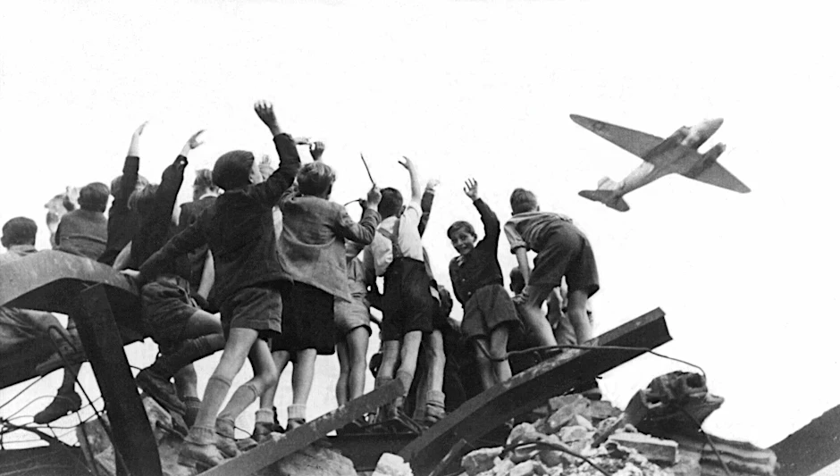 West-Berliner Jungen stehen auf einem Trümmerberg und beobachten ein amerikanisches Transportflugzeug, das Versorgungsgüter nach West-Berlin bringt.