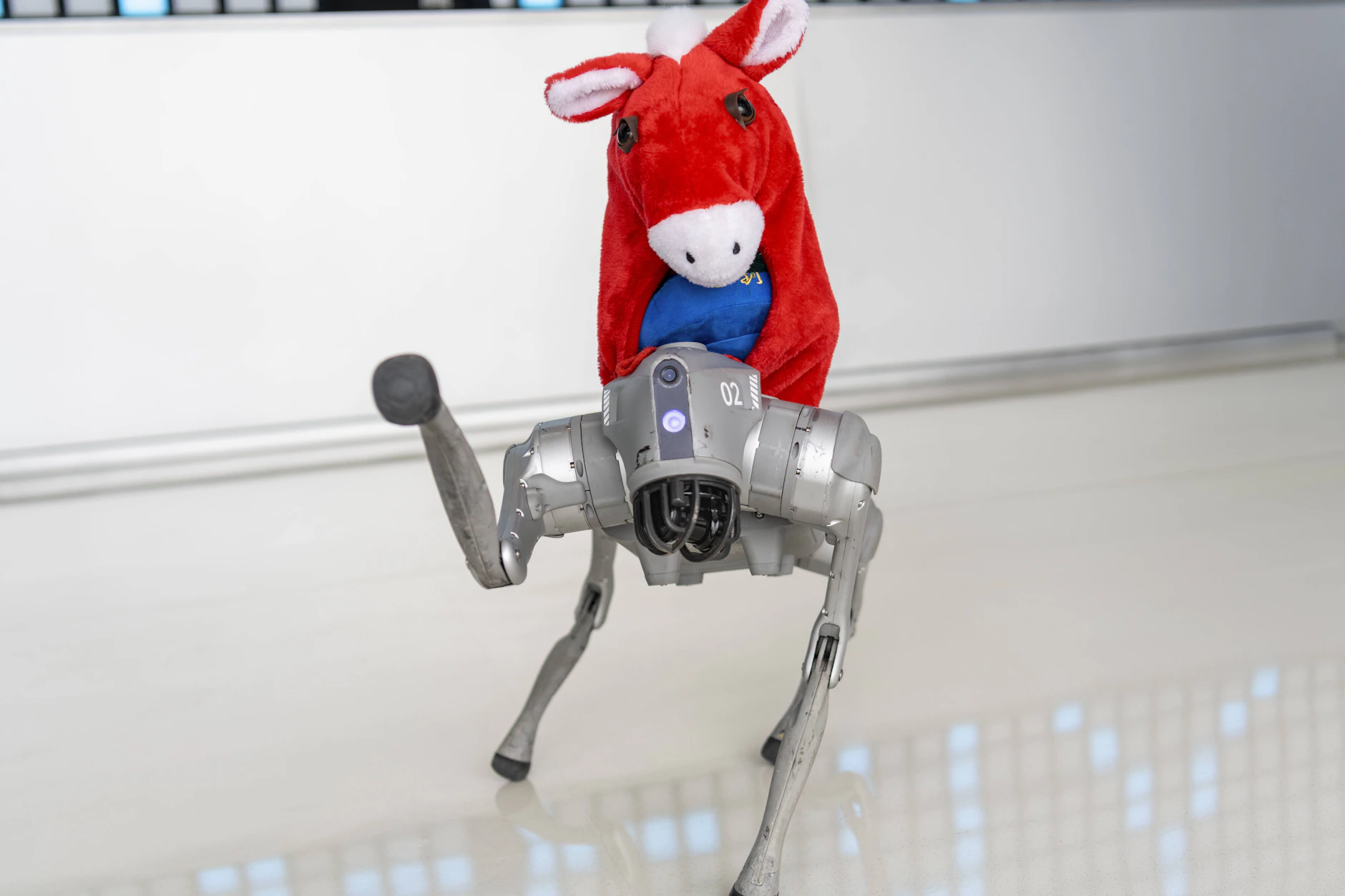Ein vierbeiniger Roboterhund aus Metall hebt ein Bein, während ein rotes Kuscheltier auf seinem Rücken sitzt. Im Verborgenen lösen Algorithmen komplexe Mathematikaufgaben auf Weltklasseniveau – doch der Einsatz modernster Maschinen wirkt in der physischen Welt oft noch unfreiwillig komisch.