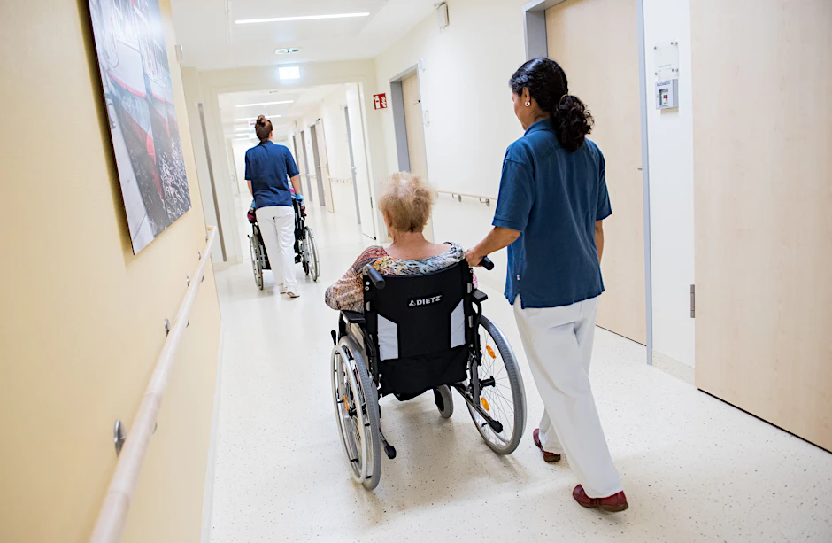 Zwei Pflegerinnen schieben in einem Klinikum Patienten mit Rollstühlen durch einen Flur.