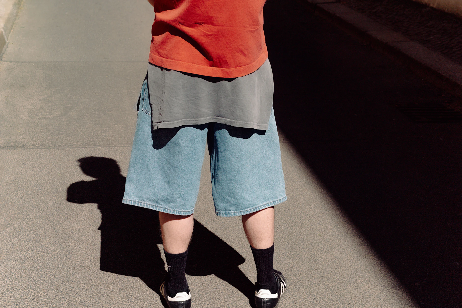 Marc setzt auf einen lässigen Skater-inspirierten Lagen-Look mit rotem T-Shirt, weiten Jeans-Shorts und Adidas Sambas. Locker um die Hüfte geknotet: ein graues Longsleeve. Seine Accessoires sind persönliche Erinnerungsstücke: Ein Silberring vom Straßenmarkt aus seinem Auslandssemester und eine Freundschaftskette mit einer Murano-Glasperle, die er sich gemeinsam mit Robinson in Venedig kaufte.