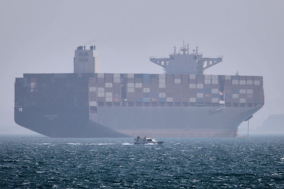 Ein Containerschiff liegt vor Anker, während im Vordergrund ein kleines Motorboot in der Straße von Hormus vor vorbeifährt.