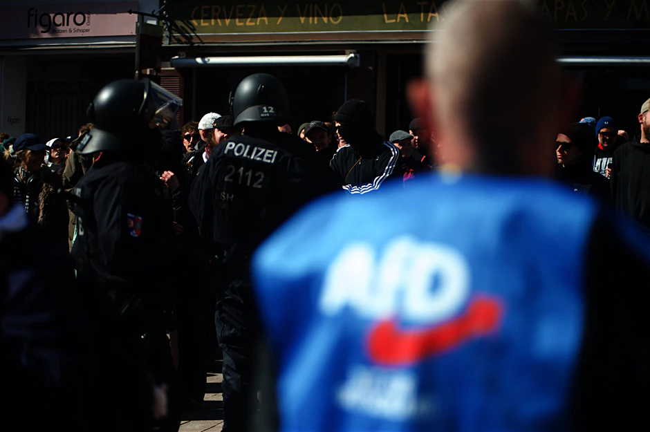 Ein AfD-Politiker beobachtet eine Szene zwischen Demonstranten und der Polizei vor einem Infostand der Partei in Flensburg.
