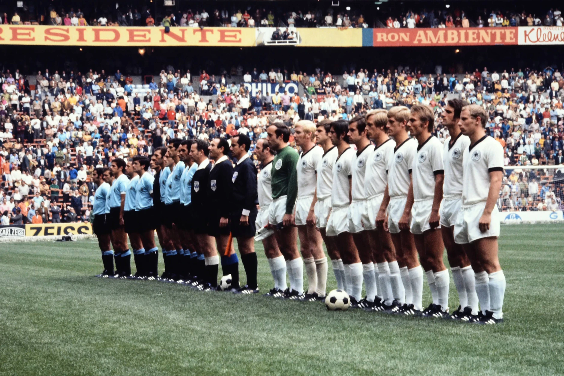 Das Spiel um Platz 3 bei der WM 1970 in Mexiko: Die Mannschaften von Deutschland und Uruguay reihen sich vor dem Anpfiff auf. Dabei sticht die Werbebande mit Carl Zeiss aus der DDR hervor.