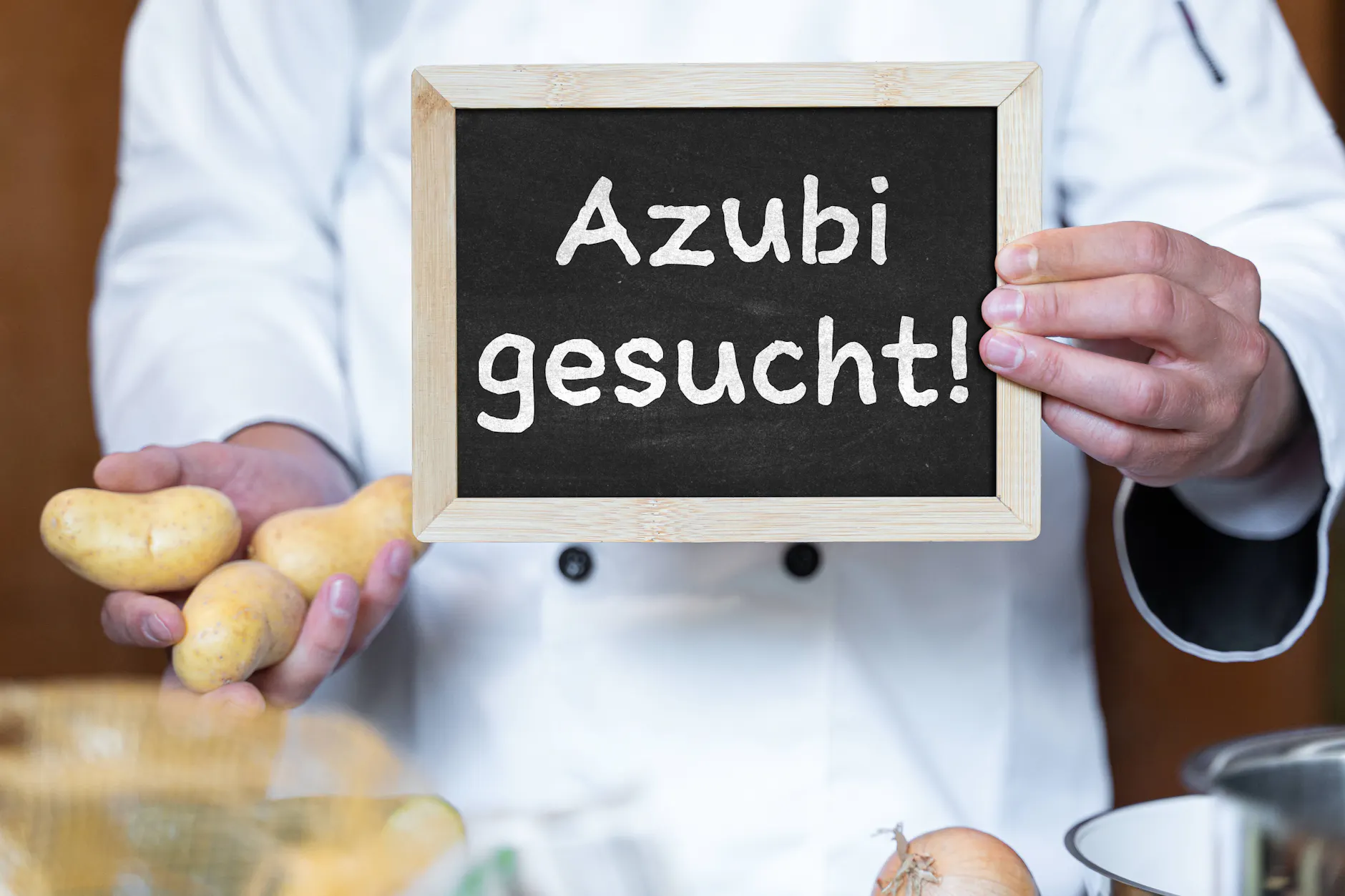 Ein Koch hält in der Küche ein Schild mit der Aufschrift: Azubi gesucht!