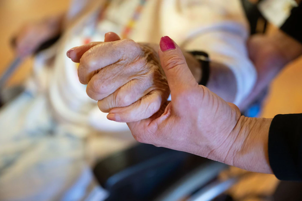 Eine Pflegekraft hält die Hand einer alten Frau – ein Bild, das zeigt, was die Reform verhandelt: menschliche Nähe, die ihren Preis hat.