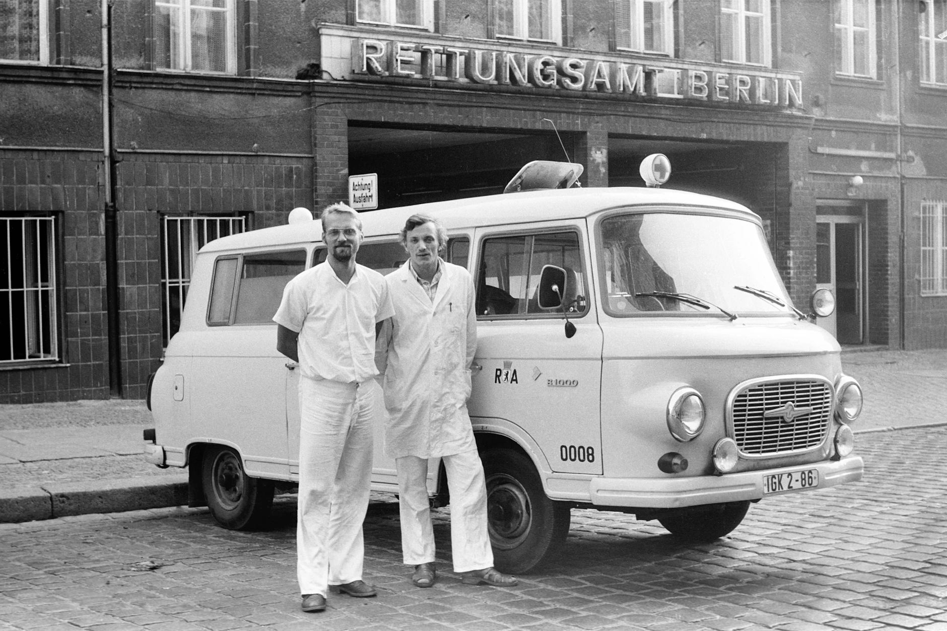 Zwei Krankentransporteure des Rettungsamtes Berlin vor ihrem Fahrzeug vom Typ Barkas B 1000. Im Hintergrund die Einfahrt zum Rettungsamt in der Marienburger Straße.