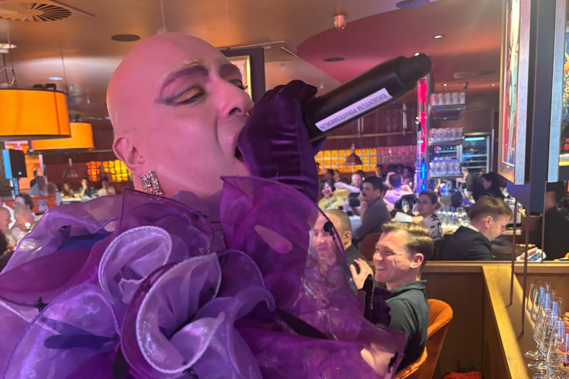 Drag-Performer in violettem Rüschenkostüm trinkt während des Auftritts aus einer Flasche, Gäste im Hintergrund beim Pink Pride Dinner Berlin