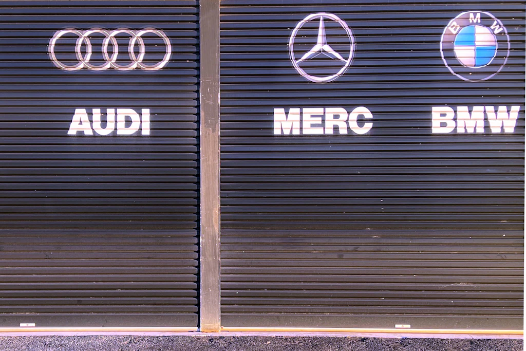 Schilder der deutschen Autohersteller Volkswagen, Audi, Mercedes-Benz und BMW an den Garagentoren einer Autowerkstatt. (Symbolbild)