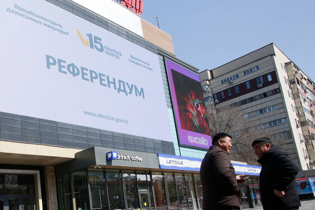 Zwei Männer stehen am 14. März 2026 in Almaty vor einer Großbildanzeige zum Verfassungsreferendum, das am folgenden Tag stattfand.