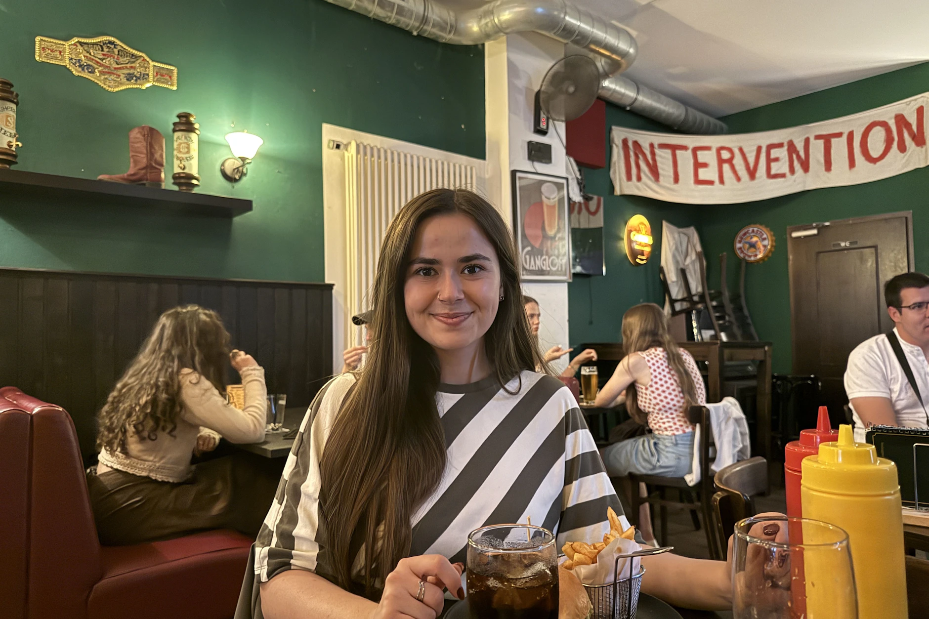 KURIER-Reporterin Mariella Mandurino verspeiste einen Lily‘s Crispy Chicken Burger. Im Hintergrund ist ein riesiges „Intervention“-Banner zu sehen. Auch das war in der Serie ein Gag.