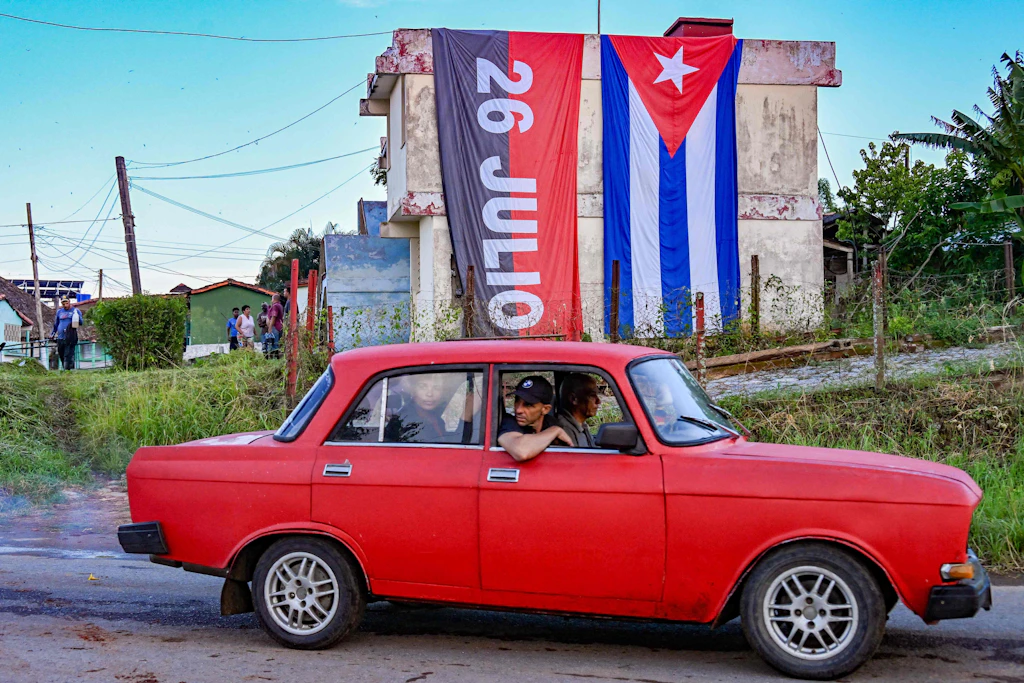 Kuba: Sowjet-Auto fährt in Pinar del Río an Haus mit Kubas und 26.-Juli-Flaggen vorbei.