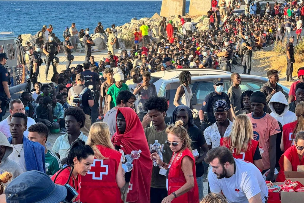 Helfer des Roten Kreuzes verteilen in Ceuta Wasser an Migranten, dahinter sichern spanische Polizisten das Ufer.