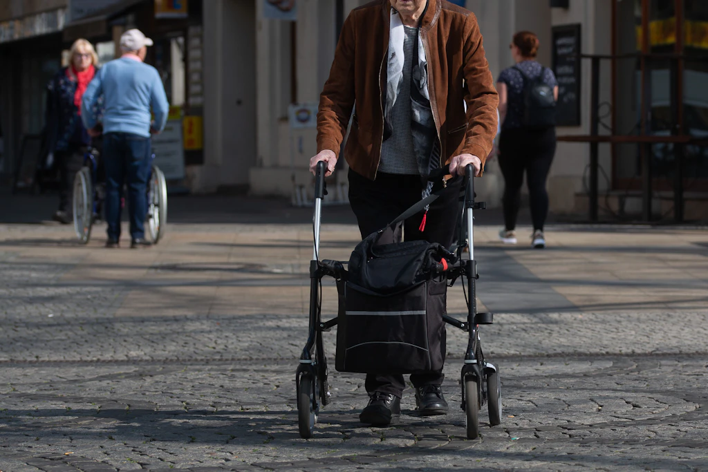 Eine ältere Person geht mit einem Rollator über einen gepflasterten Platz. (Archivbild)
