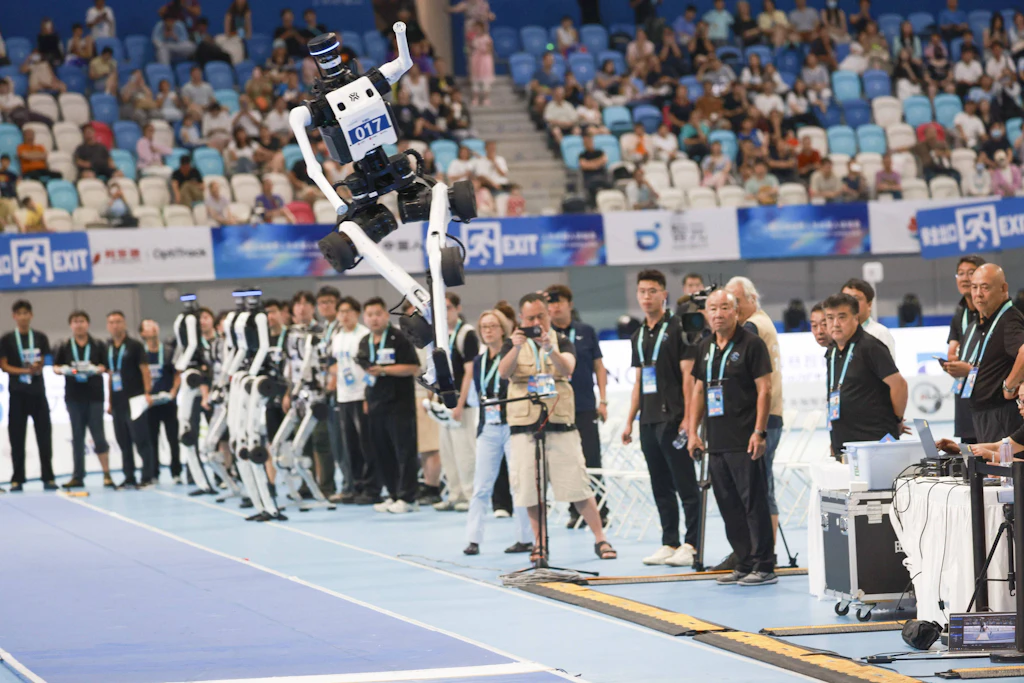 Sprungwettbewerb bei den World Humanoid Robot Games in Peking. Vor der Tribüne stehen die Konkurrenten in einer Reihe, daneben ihre Ingenieure.