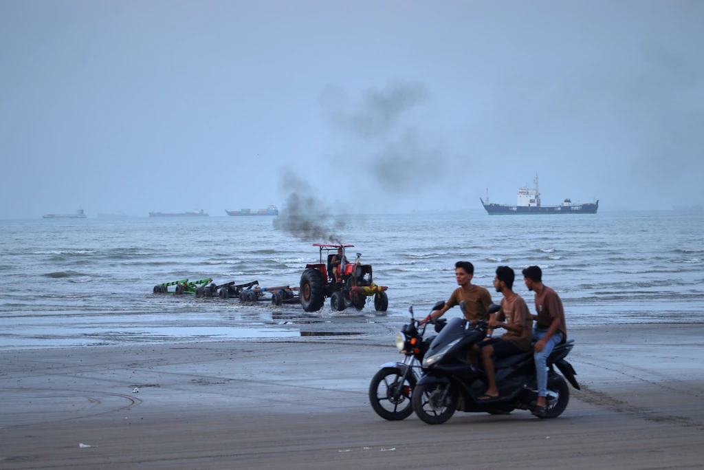 Junge Männer fahren mit Motorrädern am Ufer entlang, während ein Traktor Geräte aus dem Wasser zieht und Handelsschiffe in der Straße von Hormuz vor Anker liegen.