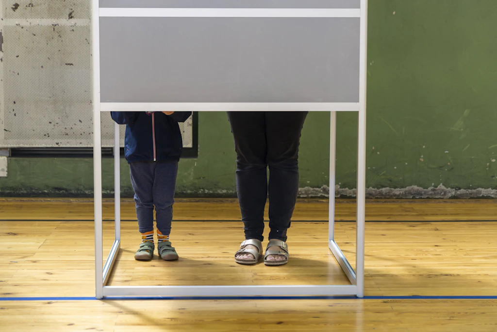 Mutter und Kind in einem Wahllokal: Nur die Mutter kann politisch mitentscheiden, was auch gut so ist.
