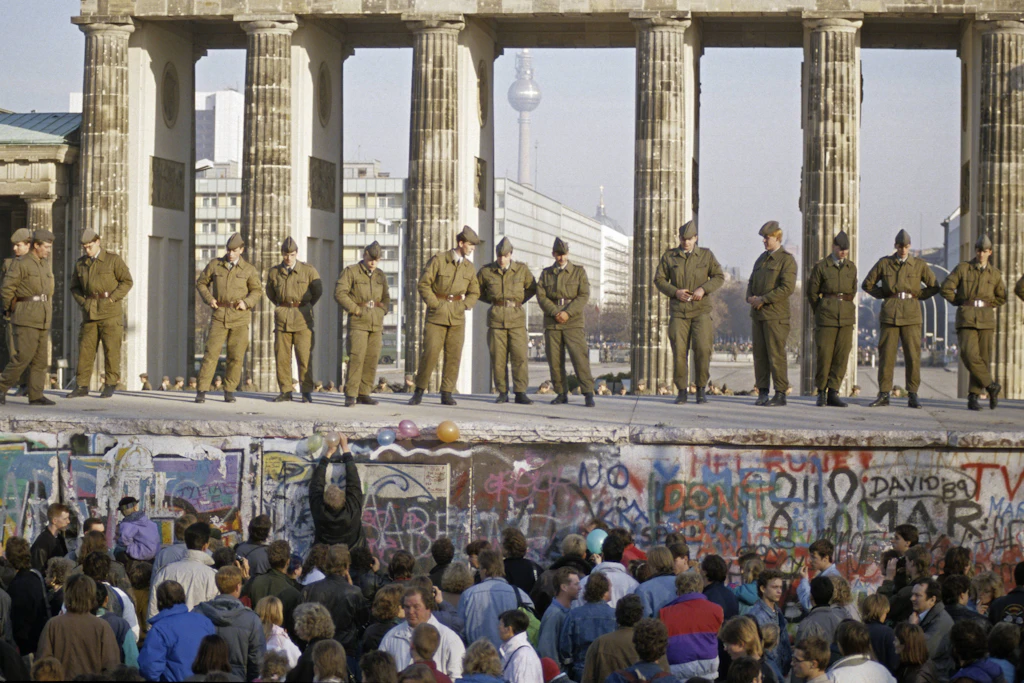 Grenzer auf der Berliner Mauer und Menschenmassen davor am Brandenburger Tor während des Mauerfalls.