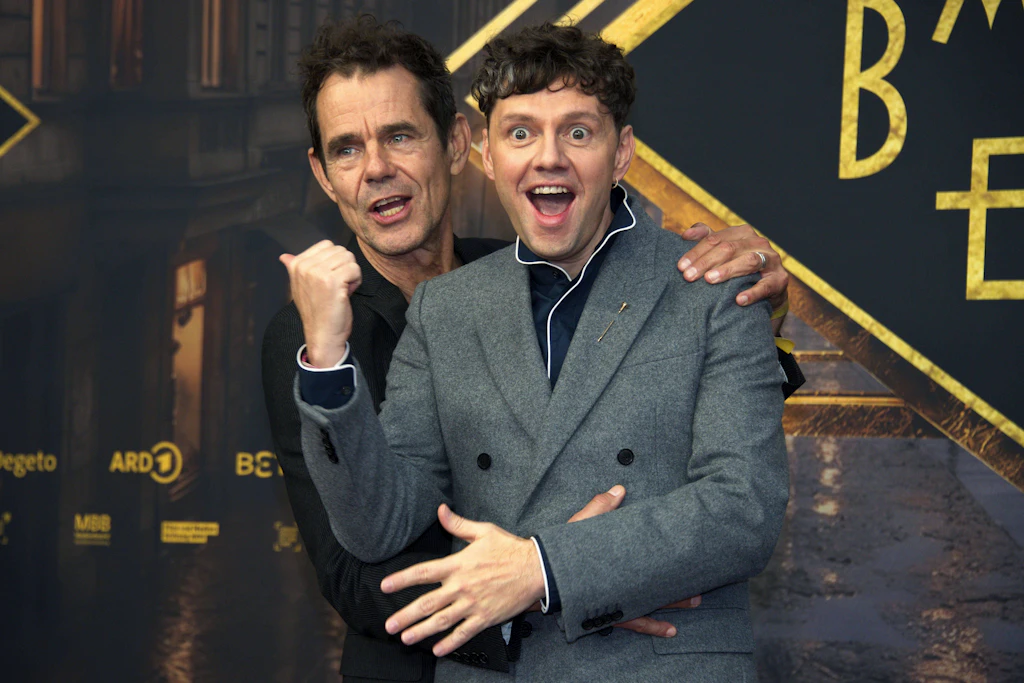 Tom Tykwer und Christian Friedel posieren mit überrascht aufgerissenen Augen vor der goldenen „Babylon Berlin“-Wand bei der Premiere im Zoo Palast.