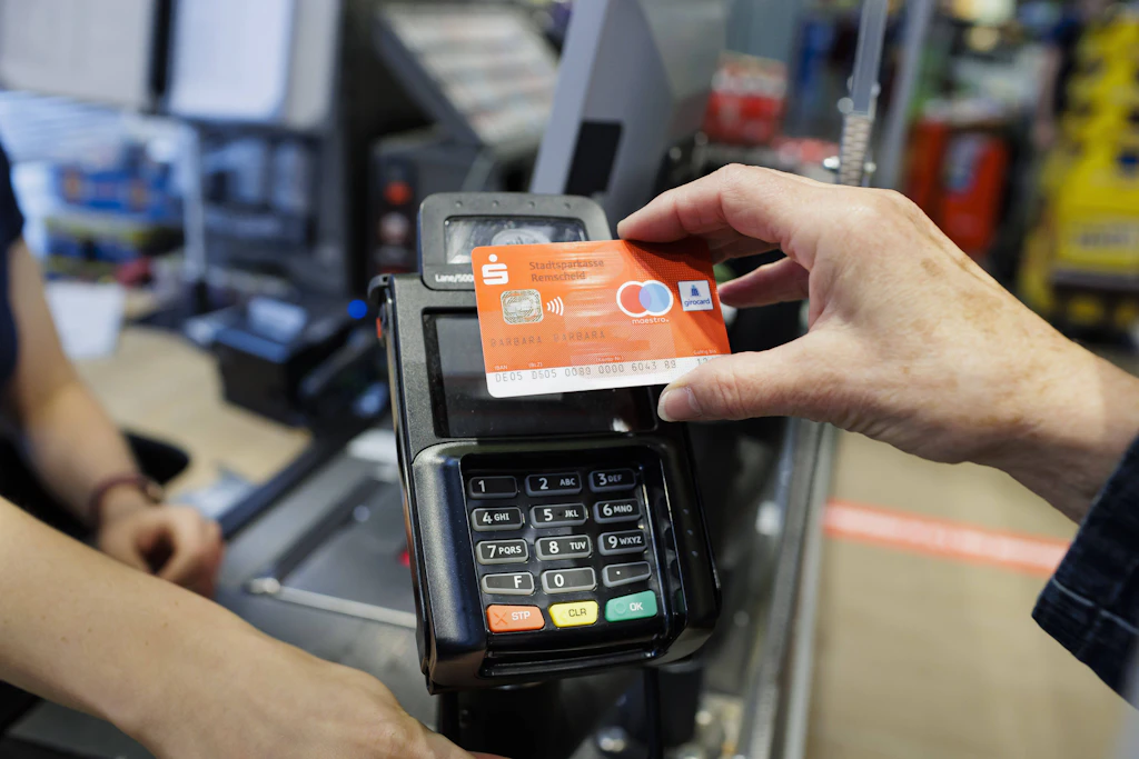 Eine Hand hält eine Girocard der Sparkasse zum kontaktlosen Bezahlen an ein Kartenterminal an einer Ladenkasse, im Hintergrund eine Kassiererin.