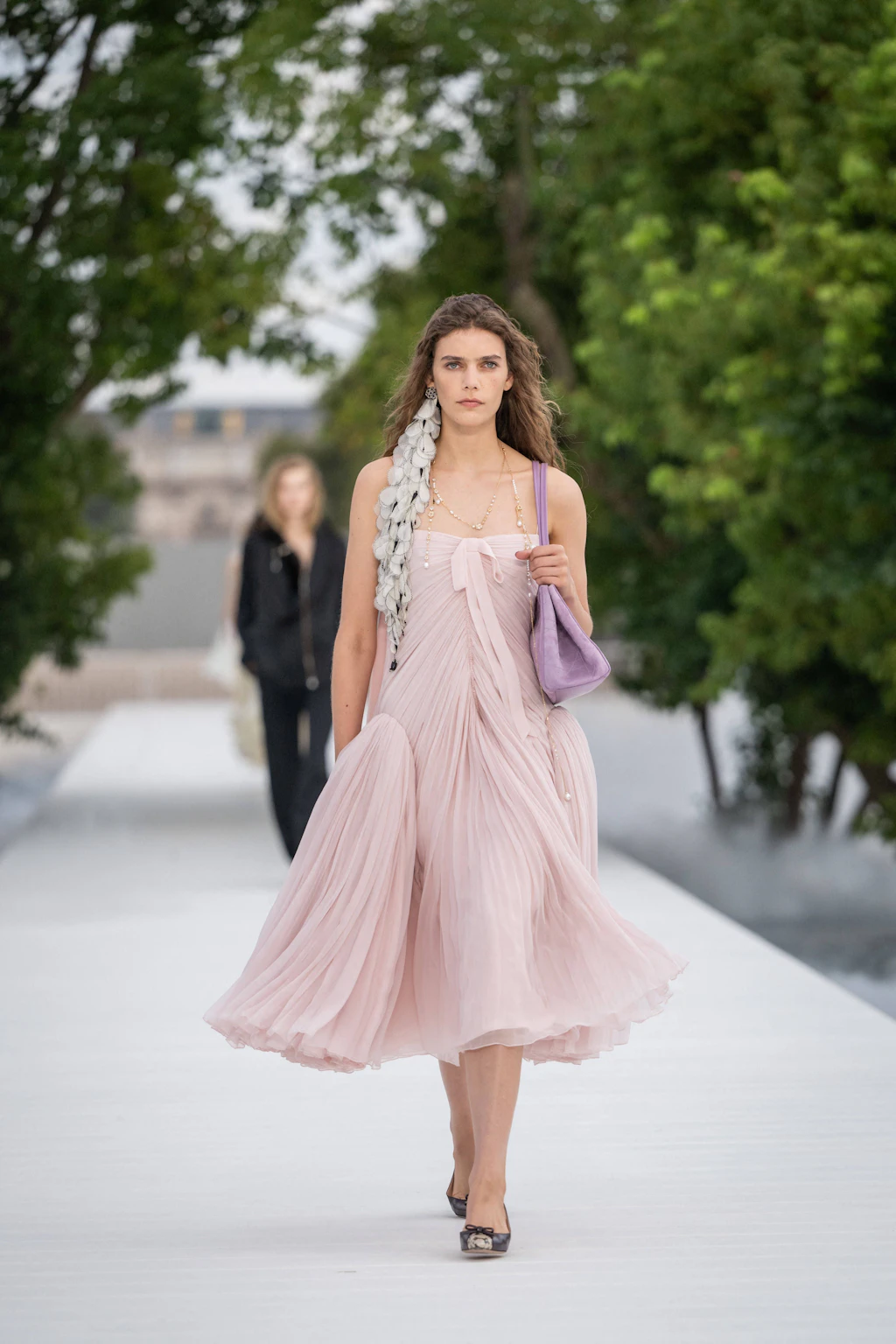 „Wie verhält sich ein Kleidungsstück im Wind?“ – Diese Frage diente dem Dior-Kreativdirektor Jonathan Anderson als Leitmotiv für die jüngste Kollektion. Neben einer Neuerfindung klassischer Dior-Stilmerkmale liegt der Fokus auf frühlingshafter Leichtigkeit, Bewegung und fließenden Silhouetten. Bei dem gezeigten Look handelt es sich um eine moderne, luftige Interpretation des ikonischen „La Cigale“-Kleides des Hauses aus dem Jahr 1952, das hier in zartem, plissiertem rosa Tüll und mit asymmetrischem Faltenwurf neu aufgelegt wurde. Die langen, floralen Ohrringe zogen sich als wiederkehrendes Motiv durch die gesamte Show.