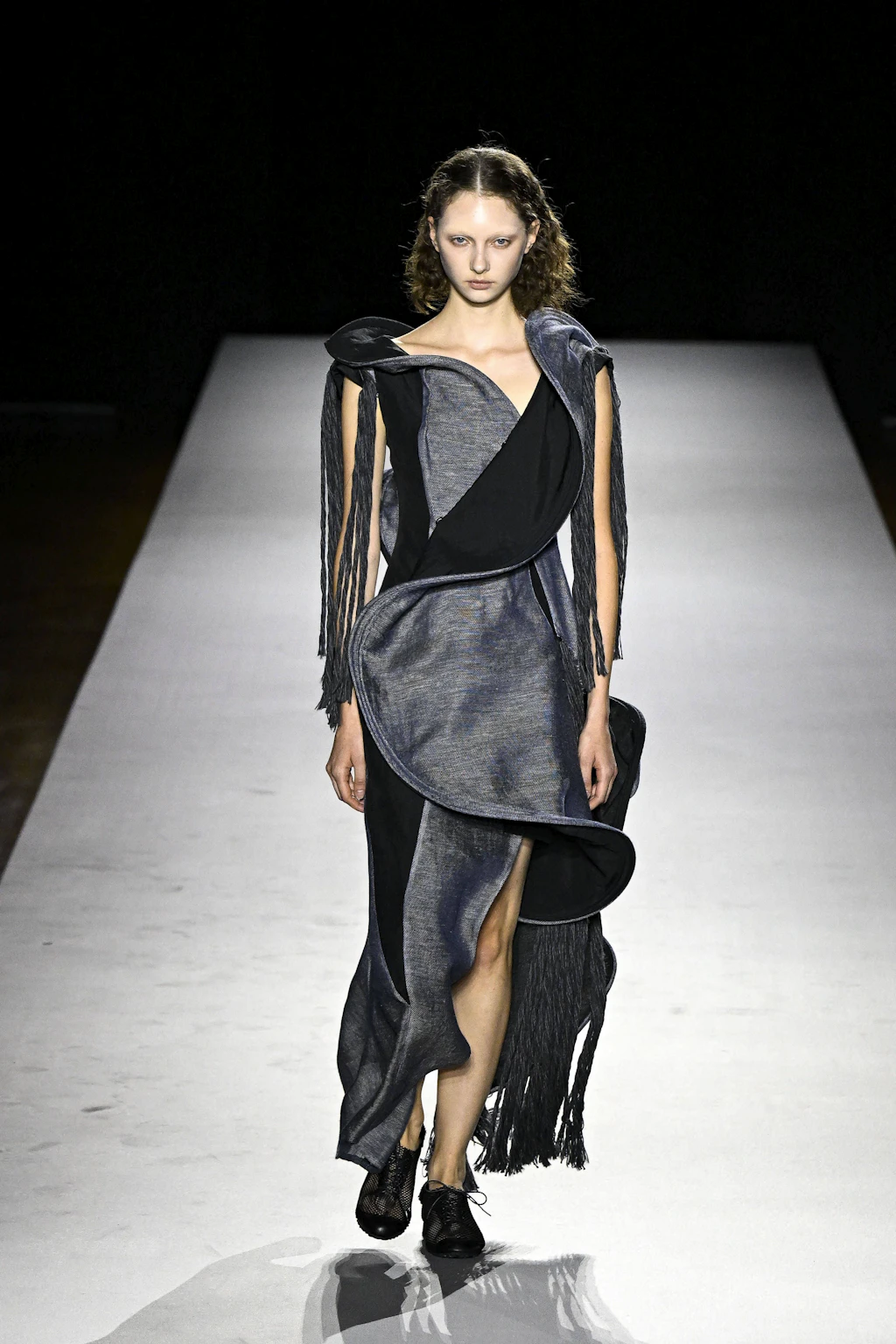 Mit der Kollektion für Frühling/Sommer 2027 treibt Yohji Yamamoto seine markante Ästhetik der Dekonstruktion auf die Spitze. Unter dem Leitmotiv „just fringes“ (nur Fransen) inszeniert der 83-jährige Designer im Grunde ein textiles Vergehen: Die Designs scheinen sich im Gehen aufzulösen. Bodenlange Fransen treffen auf Drahtkonstruktionen, die den Körper wie fragile Schutzpanzer umhüllen. Die monochrome Palette aus Schwarz, Weiß und Grau wird einzig durch einen finalen roten Look aufgebrochen. Ein melancholisches Spiel mit der Vergänglichkeit und der Poesie der Unvollständigkeit.