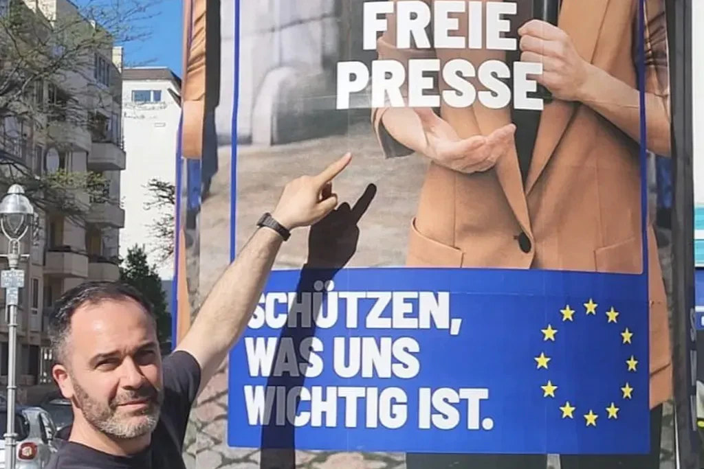 Die Kommission wirbt, der Rat sanktioniert: Hüseyin Doğru im Mai 2026 vor einem EU-Plakat, das für „freie Presse“ wirbt.