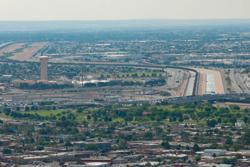 Die Grenze zwischen El Paso und Ciudad Juárez schlängelt sich durchs Bild. Unterhalb der X-förmigen Skulptur staut sich der Verkehr auf der Bridge of the Americas in Richtung USA. Zu sehen sind auch der Grenzzaun, der Rio Grande, der Border Highway und ein Bewässerungskanal.