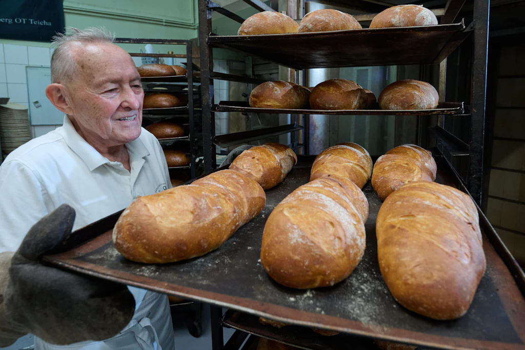 Der 85-jährige Bäckermeister Gert Ebenrecht zieht in der Backstube der Bäckerei und Konditorei in Teicha ein Blech mit frisch gebackenen Broten aus einem Wagen.