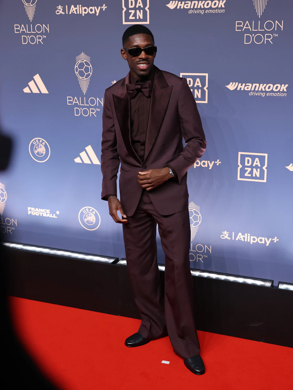 <strong>Top:</strong> Black Tie? Nicht mit Ballon-d’Or-Titelverteidiger Ousmane Dembélé. Der Spieler von Paris Saint-Germain entschied sich für einen herbstlichen Look in Burgunderrot – von Kopf bis Fuß. Sein eleganter Anzug von Zegna sitzt leicht oversized, Hemd und Fliege greifen denselben Farbton auf. Wir haben nichts auszusetzen: Mut zur Farbe zahlt sich manchmal aus, auch wenn es nur eine einzige ist. Einzig die Sonnenbrille hätte Dembélé für den roten Teppich absetzen können.